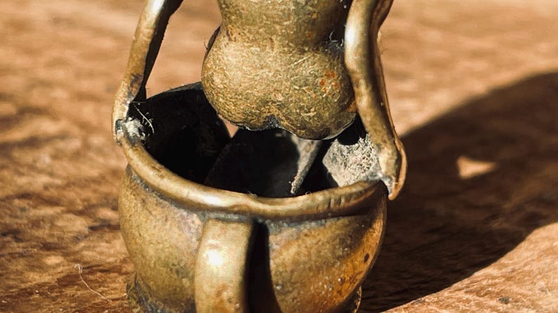 Pu&ograve; includere: Una piccola scultura in metallo dorato che rappresenta una persona seduta in una pentola. La scultura &egrave; dettagliata e ha un aspetto rustico e invecchiato.