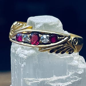 May include: A gold ring with a delicate design featuring a row of red rubies and a single diamond. The ring is set on a white background.