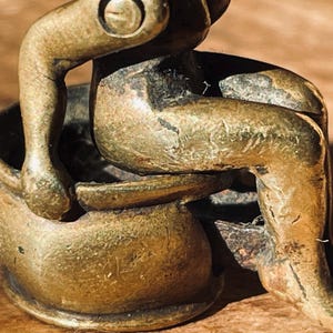 Pu&ograve; includere: Una piccola figurina in metallo dorato di una persona seduta su un vaso. La figurina &egrave; dettagliata e ha un aspetto usurato e antico.
