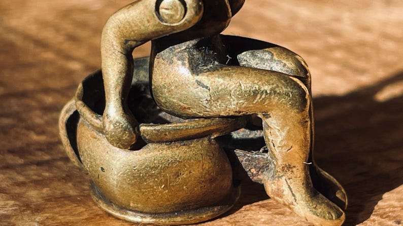 Pu&ograve; includere: Una piccola figurina in metallo dorato di una persona seduta su un vaso. La figurina &egrave; dettagliata e ha un aspetto usurato e antico.