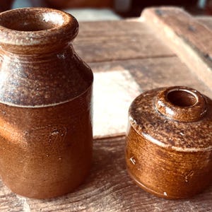 May include: Two brown ceramic inkwells, one tall and slender, the other shorter and wider. Both have a glazed finish and a slightly textured surface.