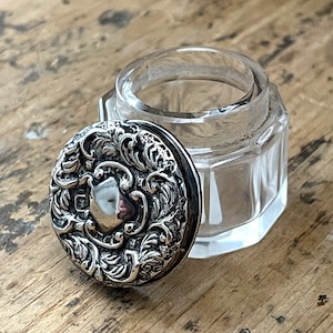 Peut inclure: Un petit pot en verre transparent avec un couvercle en argent. Le couvercle est décoré d'un motif floral complexe.