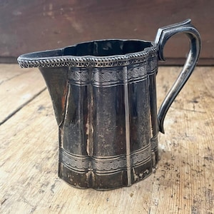May include: A silver creamer with a fluted design and a decorative border. The creamer has a handle and a spout.