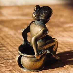 Pu&ograve; includere: Una piccola statuetta di bronzo di una persona seduta su un vaso. La statuetta &egrave; dettagliata e ha un aspetto realistico.