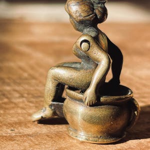 Pu&ograve; includere: Una piccola statuetta in bronzo antico di una donna seduta su un vaso. La statuetta &egrave; dettagliata e ha una finitura rustica.