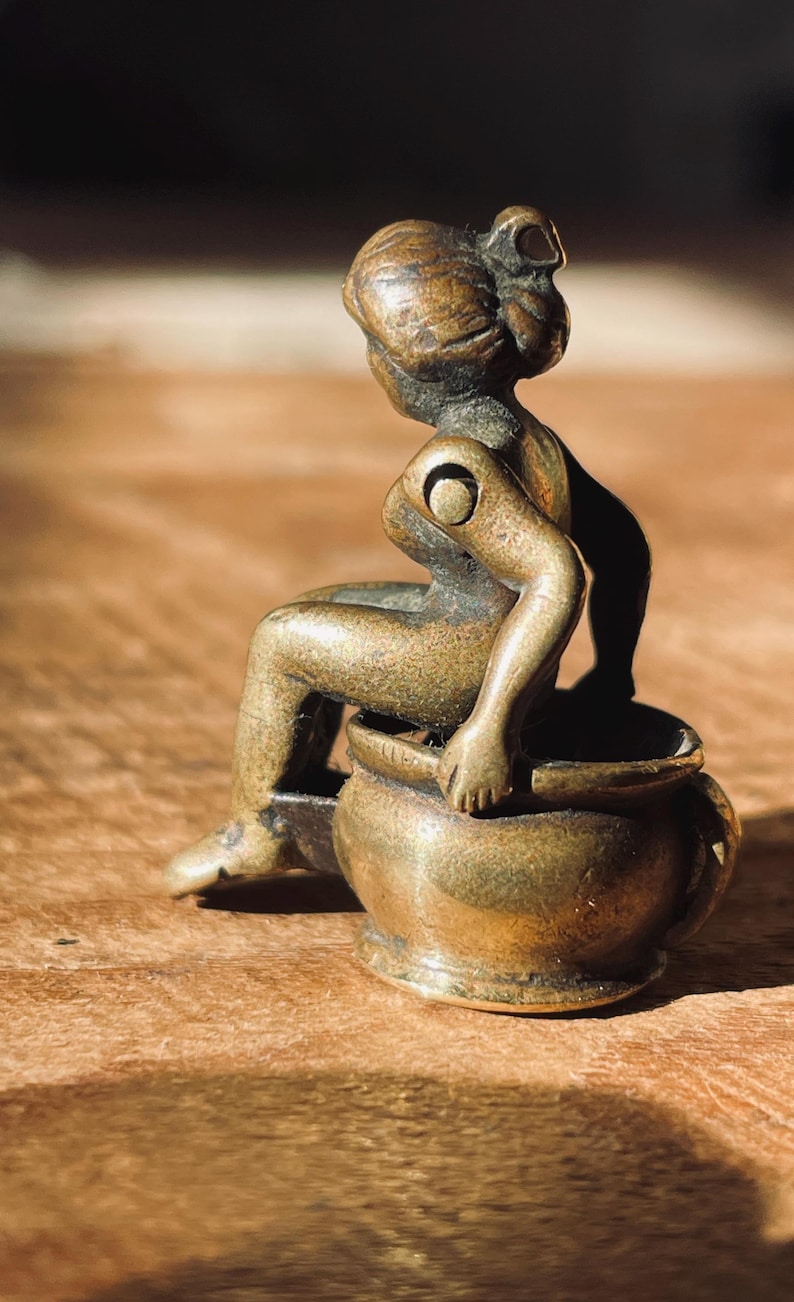 Pu&ograve; includere: Una piccola statuetta in bronzo antico di una donna seduta su un vaso. La statuetta &egrave; dettagliata e ha una finitura rustica.