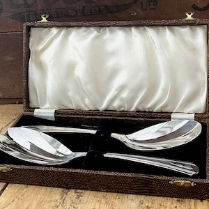 May include: A set of two silver serving spoons in a brown velvet-lined box. The spoons have a decorative design and are in excellent condition.