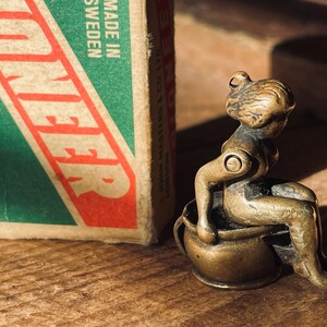 Pu&ograve; includere: Una piccola statuetta di bronzo di una persona seduta su un vaso. La statuetta &egrave; dettagliata e ha un aspetto realistico. La persona &egrave; seduta con le gambe incrociate e le braccia appoggiate sulle ginocchia. Il vaso &egrave; decorato con un semplice disegno.