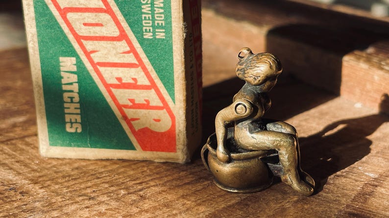 Pu&ograve; includere: Una piccola statuetta di bronzo di una persona seduta su un vaso. La statuetta &egrave; dettagliata e ha un aspetto realistico. La persona &egrave; seduta con le gambe incrociate e le braccia appoggiate sulle ginocchia. Il vaso &egrave; decorato con un semplice disegno.