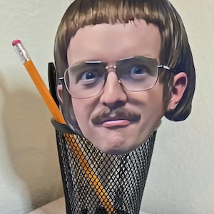 May include: A novelty pencil holder featuring a cutout of a man's face with glasses and a mustache. The holder is a black wire mesh cup, with a yellow pencil and a black pen inside.