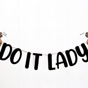 May include: A banner with the words "DO IT LADY" in bold black letters. The banner is strung with a thin cord. On each side of the banner is a cutout image of a man in orange shorts. One image has the text "On your birthday" and the other has the text "All of it".