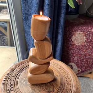 May include: A wooden candle holder with a lit candle. The holder is made of stacked, rounded wooden pieces. The candle holder sits on a carved wooden table. The background includes a blue curtain and a patterned rug.