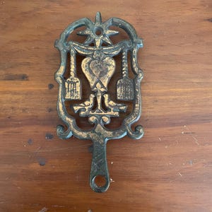 May include: A cast iron trivet with a heart and two birds design. The trivet is painted in a green and gold color scheme.