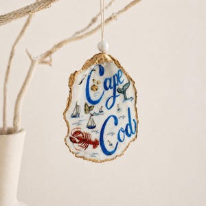 May include: A decorative oyster shell ornament with the words "Cape Cod" in blue script. The shell features painted images of a lobster, sailboats, and a whale. The shell is trimmed with gold and hangs from a white string.