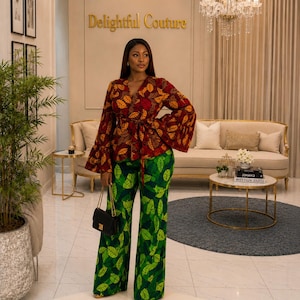 May include: A long-sleeved wrap top featuring a red, orange, and brown leaf pattern with a tie waist, paired with wide-leg trousers in a vibrant green leaf print. A small black rectangular handbag with a gold chain strap is held at the side.