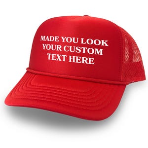 May include: Red trucker hat with a white mesh back. The front of the hat has white text that reads "MADE YOU LOOK YOUR CUSTOM TEXT HERE".