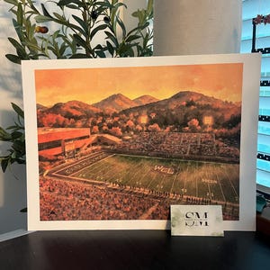 May include: A print of a football stadium at sunset. The artwork depicts a stadium with the words "CATAMOUNTS" and "WCU" visible on the field. The sky is a warm orange and yellow, with mountains in the background.