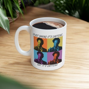 May include: White ceramic coffee mug with a black handle. The mug has a pop art design featuring four images of a man with a beard and glasses. The text on the mug reads "I'm not saying it's caffeine... but it's caffeine."