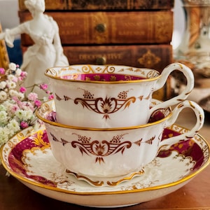 May include: Two stacked white teacups with gold trim and a matching saucer. The cups and saucer feature a burgundy and gold floral design. The teacups have curved handles. The background includes antique books and a decorative statue.