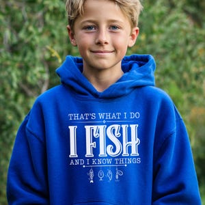 May include: A royal blue hooded sweatshirt with the text "THAT'S WHAT I DO I FISH AND I KNOW THINGS" in white. Below the text are small fishing-related graphics. The sweatshirt is worn by a person.