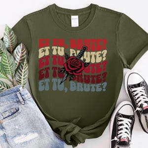 May include: A green t-shirt with a red, white, and blue graphic print of a rose and a dagger. The text "Et Tu, Brute?" is repeated in a retro font style.