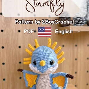 May include: A crocheted blue and cream-colored dragon with yellow accents. The dragon has large black eyes, a yellow nose, and yellow spikes on its head. The image includes the text "Stormfly" and "Pattern by 2BoyCrochet".