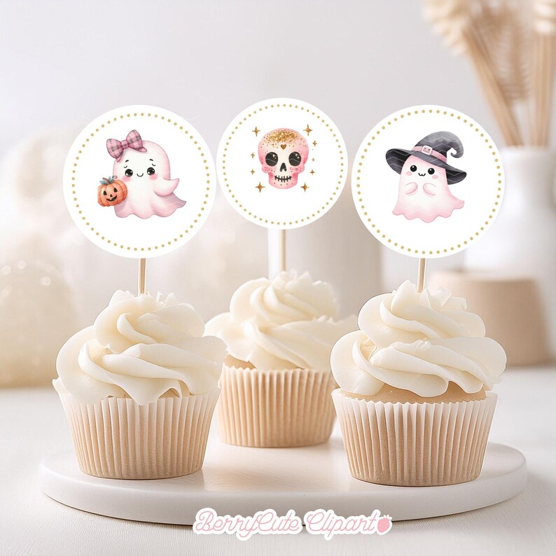 Op de afbeelding: Drie cupcake-toppers met een Halloween-thema. De toppers bevatten een spook met een roze strik, een schedel met roze en gouden details en een spook met een heksenhoed. De toppers zijn op witte cupcakes met witte frosting.