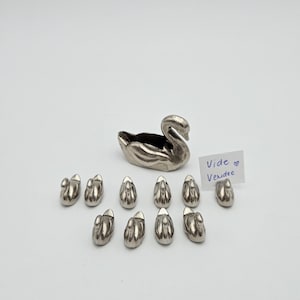 Op de afbeelding: Een zilveren zwaanvormige container met een kleine opening, vergezeld van elf miniatuur zwaanbeeldjes. Een handgeschreven notitie luidt "Vide Vendee". De objecten zijn op een wit oppervlak geplaatst.