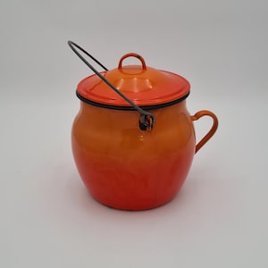 Op de afbeelding: Een vintage oranje en rode emaille pot met een deksel en een handvat. De pot heeft een zwarte rand en een draadhandvat. Hij heeft een klein handvat aan de zijkant.