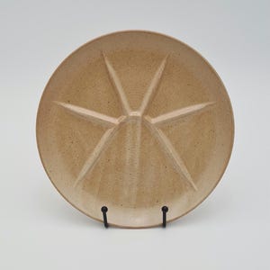 May include: A round, tan-colored ceramic plate with a starburst design. The plate is divided into six sections by raised lines. It is displayed on a black metal stand against a white background. This plate is suitable for serving appetizers or snacks.