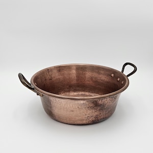 May include: A round, hammered copper basin featuring a wide, open top and two dark, curved metal handles attached to the sides with rivets. The exterior surface displays a textured, metallic finish with visible signs of natural oxidation.