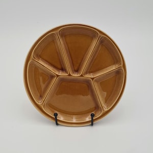 Può includere: Un piatto da portata diviso in ceramica marrone con sei scomparti. Il piatto rotondo ha un bordo rialzato e poggia su un supporto in metallo nero. Gli scomparti sono triangolari e disposti attorno a un cerchio centrale. Diametro di circa 25 cm.
