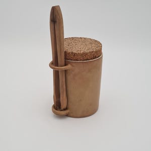 May include: A small, cylindrical, tan-colored container with a cork lid. A wooden utensil is attached to the side with a metal ring. The container is likely for storing spices or other small items.