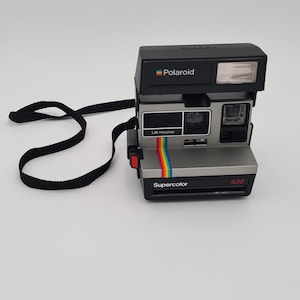 Op de afbeelding: Een vintage Polaroid Supercolor 635 camera met een zwarte behuizing en zilveren accenten. De camera heeft een ingebouwde flitser en een regenboogstreep. Het woord "Polaroid" staat erop.