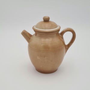 May include: A small, tan ceramic teapot with a lid and handle. The teapot has a rounded body, a spout, and a small knob on the lid. The teapot is likely used for brewing tea.