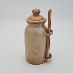 May include: A tan ceramic jar with a lid and a wooden spoon. The jar has a cylindrical shape with a slightly textured surface. The spoon is held in place by a loop on the side of the jar. The jar is approximately 5 inches tall.
