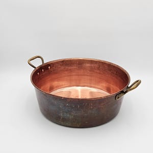 May include: A round copper cooking pot with a polished interior and a weathered, darkened exterior finish. The vessel features two curved brass handles securely attached to the sides with visible rivets, providing a sturdy grip for handling.