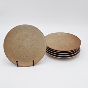 Op de afbeelding: Een set van zes ronde keramische borden met een gespikkelde beige en bruine afwerking. Eén bord staat rechtop op een zwarte metalen standaard, terwijl de overige vijf borden er netjes achter zijn gestapeld, wat hun ronde vorm benadrukt.