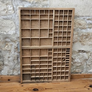 May include: A rectangular wooden printer's tray featuring a grid of numerous small, open compartments of varying sizes. The light brown wood has a natural, unfinished texture, and the structure is divided into two main horizontal sections.