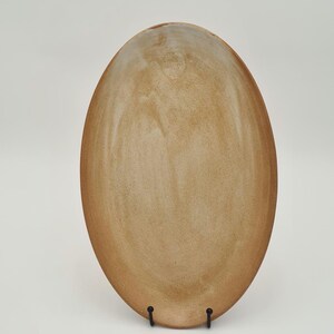 Op de afbeelding: Een grote, ovale keramische schaal in tinten bruin en beige, op een zwarte metalen standaard. De schaal heeft een glad, licht getextureerd oppervlak. Het ontwerp is eenvoudig en elegant, geschikt voor het serveren van voedsel, ongeveer 30 cm in diameter.