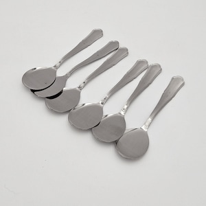 Può includere: Un set di cinque cucchiai in metallo color argento con coppe larghe e arrotondate e manici sottili e affusolati. Ogni cucchiaio presenta una finitura liscia e lucida e un design classico semplice con un'estremità leggermente svasata.