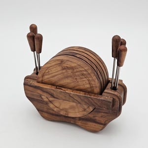Op de afbeelding: Een set ronde houten onderzetters opgeborgen in een bijpassende houten houder. De houder heeft aan beide zijden geïntegreerde sleuven, elk met drie kleine metalen aperitiefprikkers met houten handvatten. Het hout vertoont een natuurlijke nerf.
