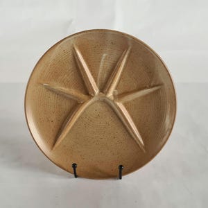 May include: A tan ceramic plate with a starburst design, divided into five sections. The plate has a speckled texture and is displayed on a black metal stand. Ideal for serving appetizers or small portions.