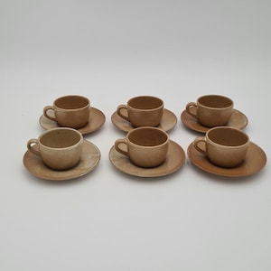 Op de afbeelding: Zes bijpassende bruine keramische espressokopjes en schotels zijn gerangschikt op een witte ondergrond. De kopjes hebben een afgeronde vorm met een klein handvat en de schotels zijn cirkelvormig. De set lijkt handgemaakt.
