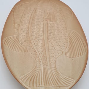 May include: Tan ceramic serving plate with a raised relief design of a fish. The fish is centered on the oval plate, with detailed scales and fins. The plate has a slightly irregular edge.