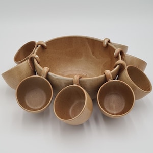 May include: A tan ceramic serving bowl with attached handles, surrounded by seven matching cups. The bowl and cups have a speckled texture and a simple, elegant design, perfect for serving snacks or dips.