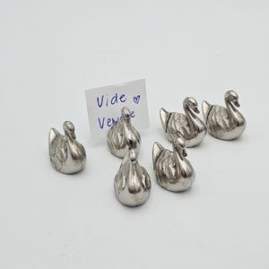 Op de afbeelding: Een verzameling van zes kleine, zilverkleurige zwaanbeeldjes. Elke zwaan heeft gedetailleerde veertjes en een gebogen nek. Er is ook een handgeschreven notitie met de woorden "Vide Vente".