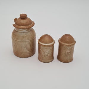 May include: A set of three ceramic kitchen containers in a light brown color. The set includes a lidded jar and two smaller shakers, one marked with an "S" and the other with a "P".