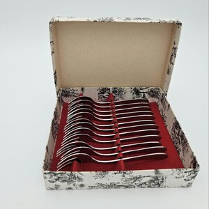 May include: A set of silver-colored spoons in a decorative box. The box has a black and white toile pattern on the exterior and a red interior. The spoons are arranged neatly in the box, suggesting a formal dining set.