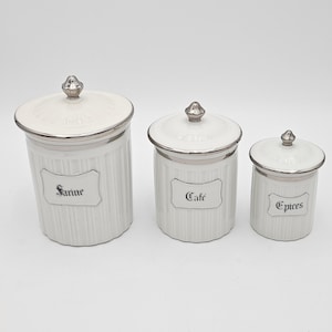 Peut inclure: Un ensemble de trois boîtes de conservation en céramique blanche avec des textures côtelées verticales et des couvercles argentés. Chaque boîte porte une étiquette décorative avec du texte noir : Farine, Café et Épices sur les trois tailles.